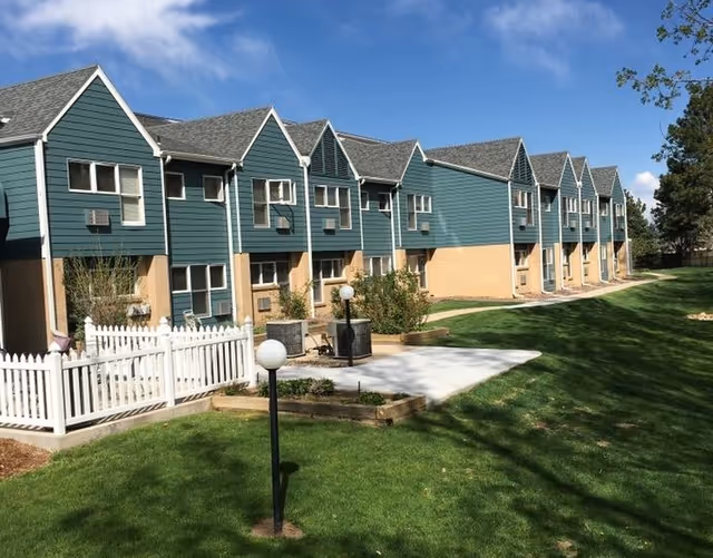 Two-story teal-and-beige apartment-style building with gabled roofs overlooking a manicured lawn, white picket fence, pathway and outdoor lamp posts.