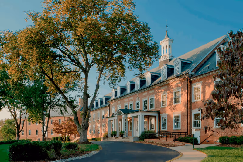 Exterior view of a large brick building with multiple windows and a cupola on the roof, surrounded by trees and greenery under a clear blue sky.
