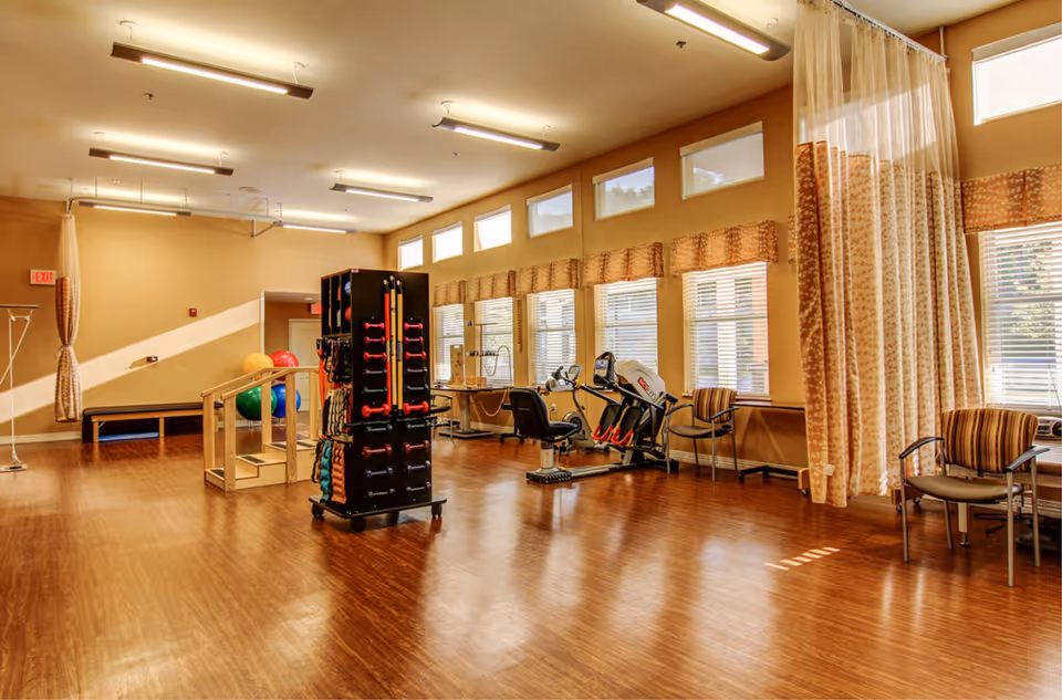 A spacious rehabilitation or physical therapy room with wooden flooring, exercise equipment including hand weights, therapy balls, parallel bars for walking practice, and a stationary exercise bike. The room has large windows with blinds and patterned curtains, several chairs along the walls, and bright overhead lighting.