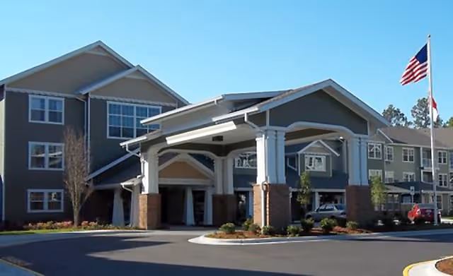 Exterior view of a multi-story senior living facility with a covered entrance, landscaped surroundings, and an American flag on a flagpole under a clear blue sky.