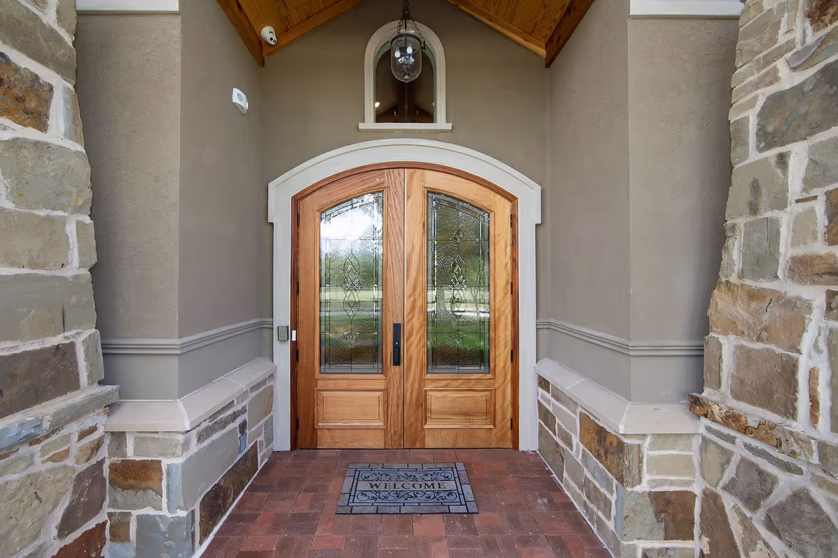 Entrance to a building with double wooden doors featuring decorative glass panels, stone walls on either side, a brick floor, and a welcome mat in front of the doors. There is a small arched window above the doors and a hanging light fixture.