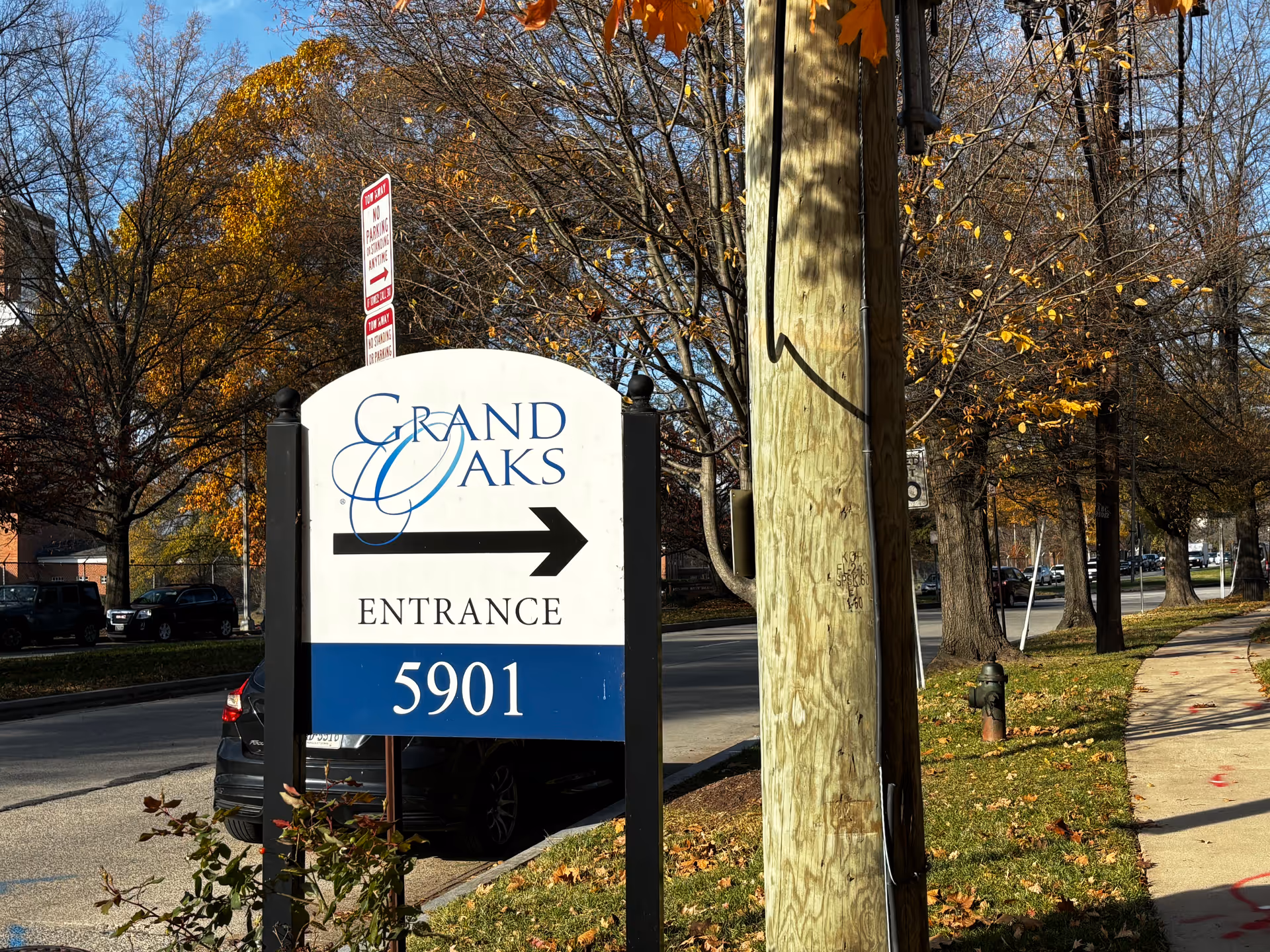 Outdoor view of a street with a sign for Grand Oaks Assisted Living Community entrance at 5901. The sign is white with blue and black text and an arrow pointing right. Trees with autumn leaves and parked cars are visible in the background.