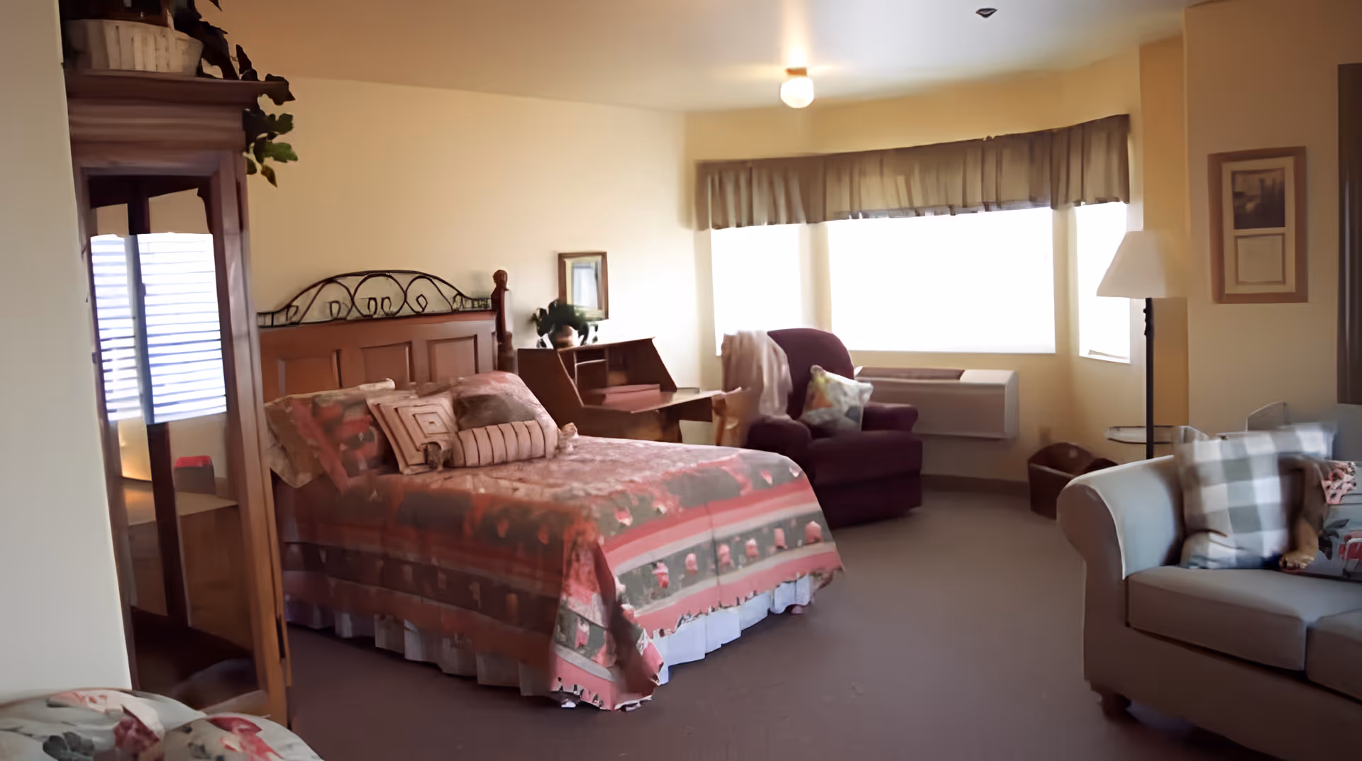 A cozy bedroom in an assisted living facility featuring a bed with a patterned bedspread and multiple pillows. The room includes a wooden dresser with a mirror, a small desk with a plant, a purple armchair with a throw blanket and pillow, a floor lamp, and a light gray sofa with checkered and floral pillows. Large windows with valances allow natural light to fill the room.