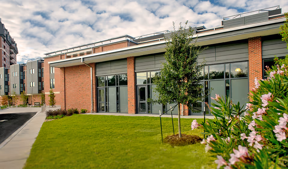 Exterior view of a modern brick and gray panel building with large windows, a green lawn, a young tree, and flowering bushes under a partly cloudy sky.