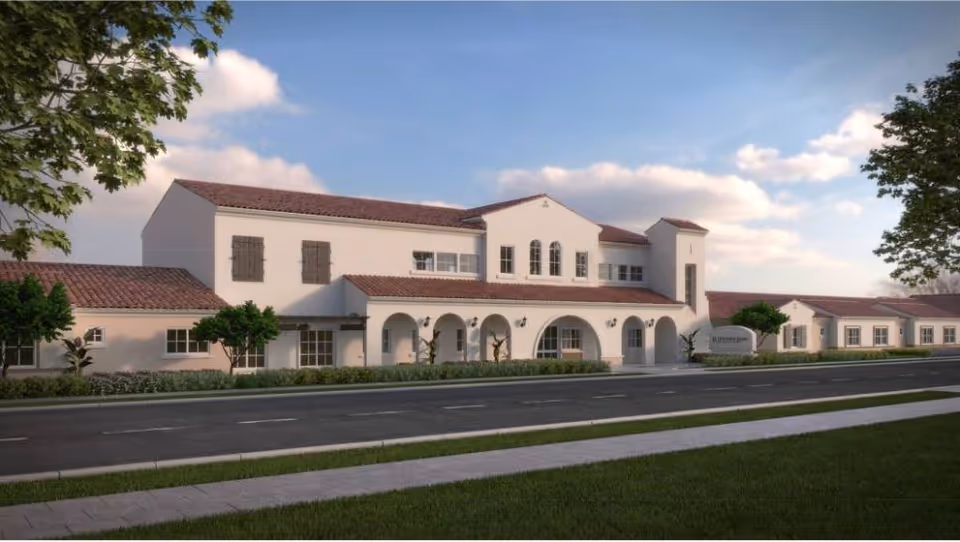 Exterior view of a large, two-story building with white walls and a red-tiled roof, featuring arched doorways and windows, surrounded by greenery and a clear sky with some clouds.