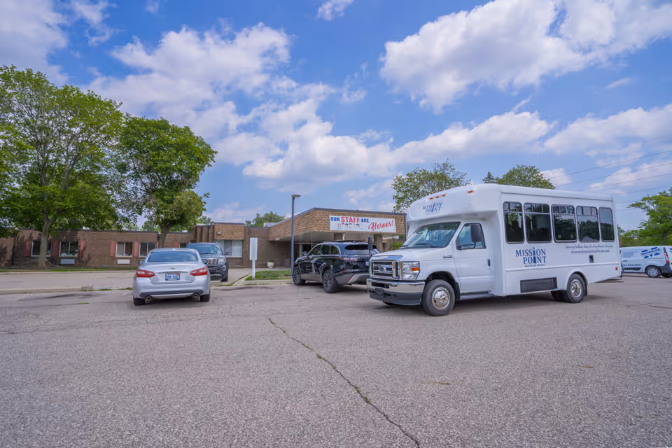 Parking lot outside a single-story brick building under a partly cloudy sky. Several vehicles are parked, including a white shuttle bus with 'Mission Point' branding on the side. A banner on the building reads 'Our Staff Are Heroes!'