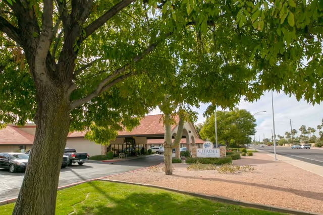 Exterior view of Citadel Post Acute facility with a large tree in the foreground, a driveway, parked cars, and a sign displaying the facility name near the entrance. The building has a tiled roof and is surrounded by landscaping with grass, bushes, and desert-style gravel.