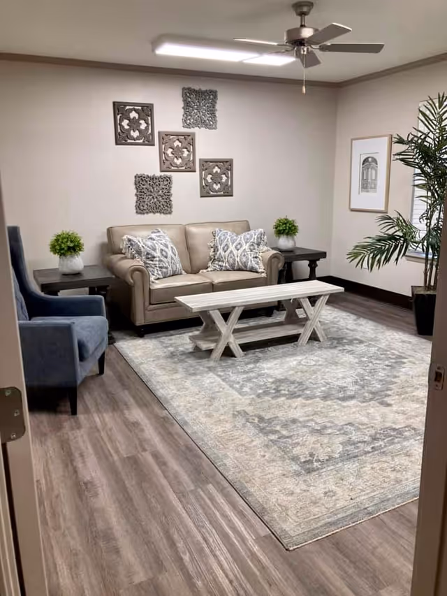 A cozy living room with a beige sofa adorned with patterned pillows, a blue armchair, a wooden coffee table, and two side tables each with a small green plant. The room features a large patterned area rug, wall decorations above the sofa, a framed picture on the wall, a ceiling fan with light, and a tall potted plant near the window.