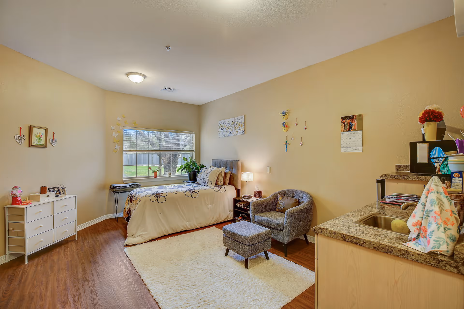 Studio bedroom in a senior living unit featuring a bed by a window, a chair and ottoman, a dresser, and a small kitchenette.