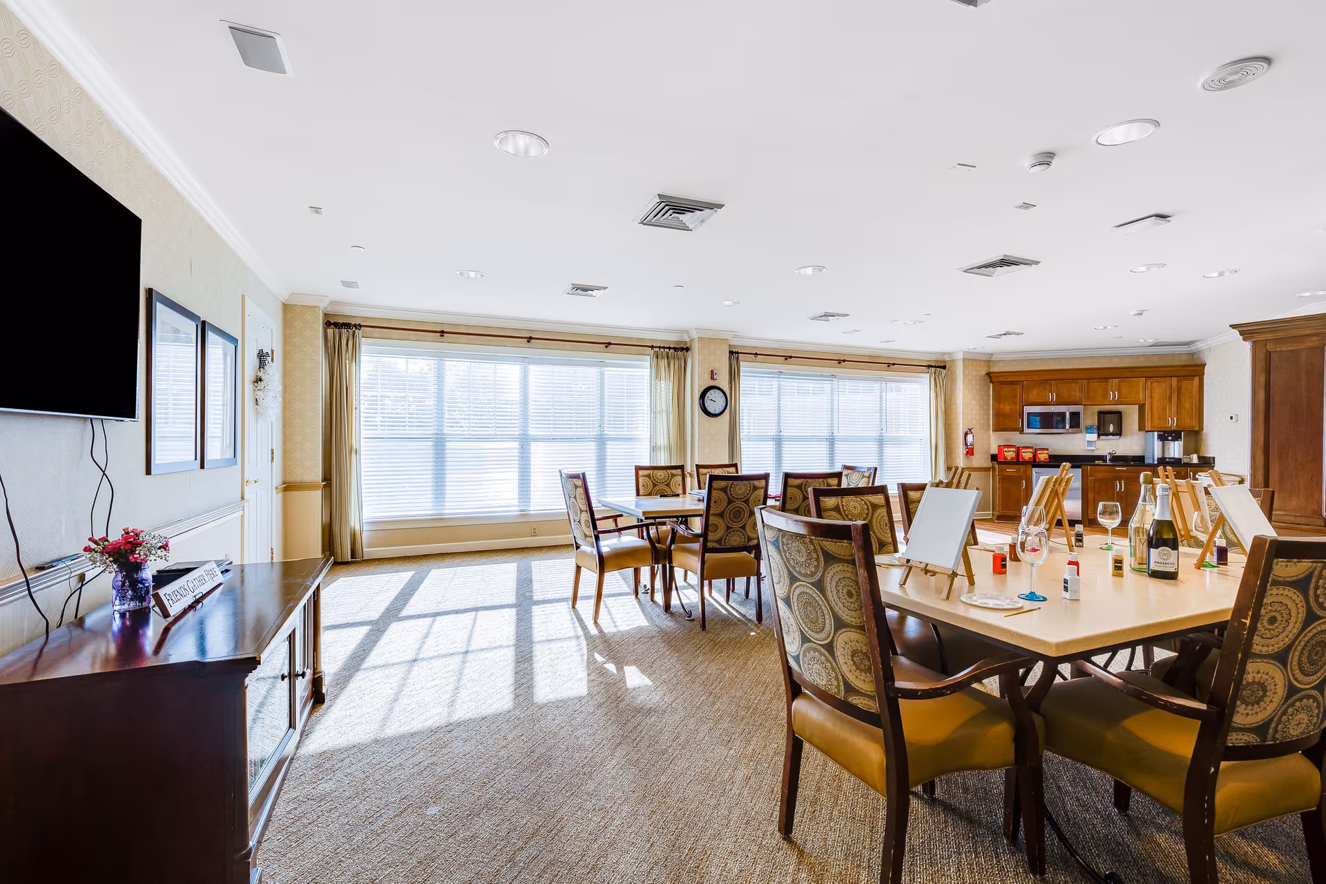 A bright and spacious common room with large windows letting in natural light. The room features multiple tables with patterned chairs arranged around them. On one table, there are art easels, paint bottles, and wine glasses, suggesting an arts and crafts activity. The back of the room has a kitchenette area with wooden cabinets, a microwave, and a coffee maker. A TV is mounted on the wall to the left, and a small cabinet with a flower vase and a sign is below it.