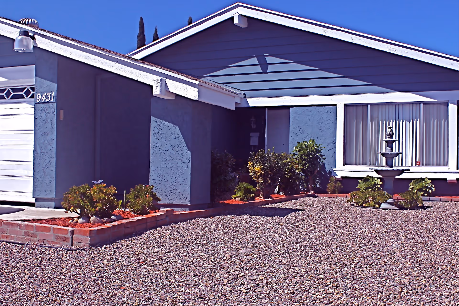 Exterior view of a single-story residential building with a gravel front yard, small garden beds with plants, a decorative fountain, and a garage door with the number 9431. The building has a blue-gray exterior with white trim and a large window with vertical blinds.