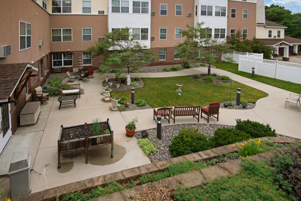Courtyard of a senior living facility with paved walkways, benches, planters, and a grassy central area in front of a multi-story building.