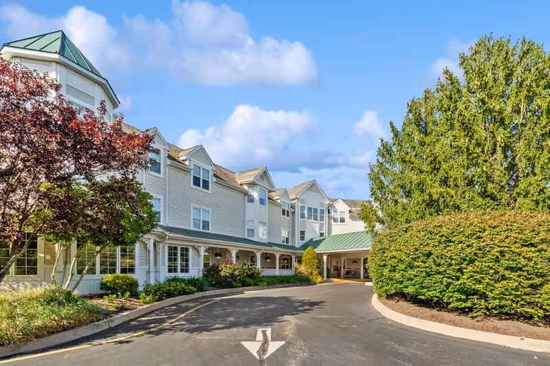 Exterior view of Woodbridge Place Senior Living facility showing a multi-story building with white siding, multiple windows, a green roofed entrance, and surrounding trees and shrubs under a partly cloudy sky.