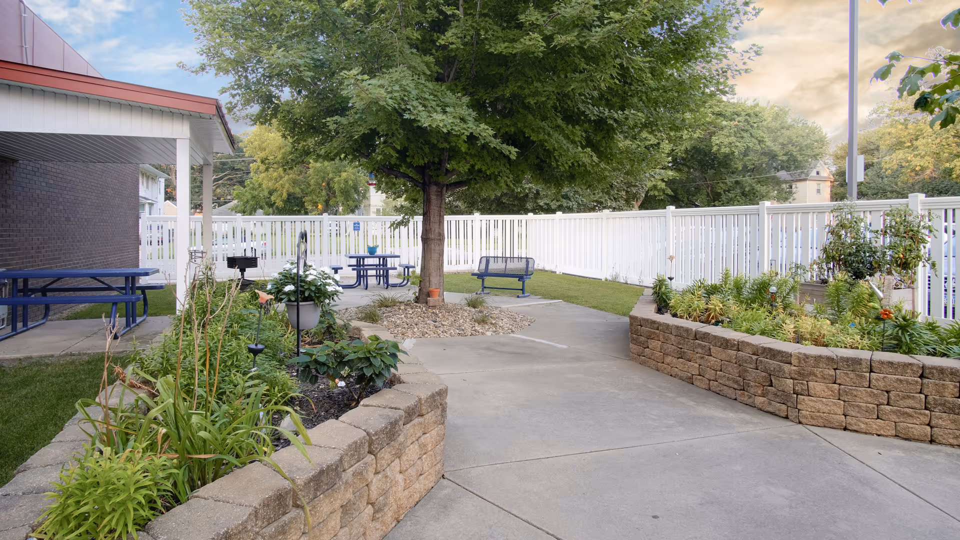 Outdoor patio area at Bridges Senior Living featuring raised garden beds with various plants, a large tree in the center, blue picnic tables, a bench swing, and a white fence surrounding the space.