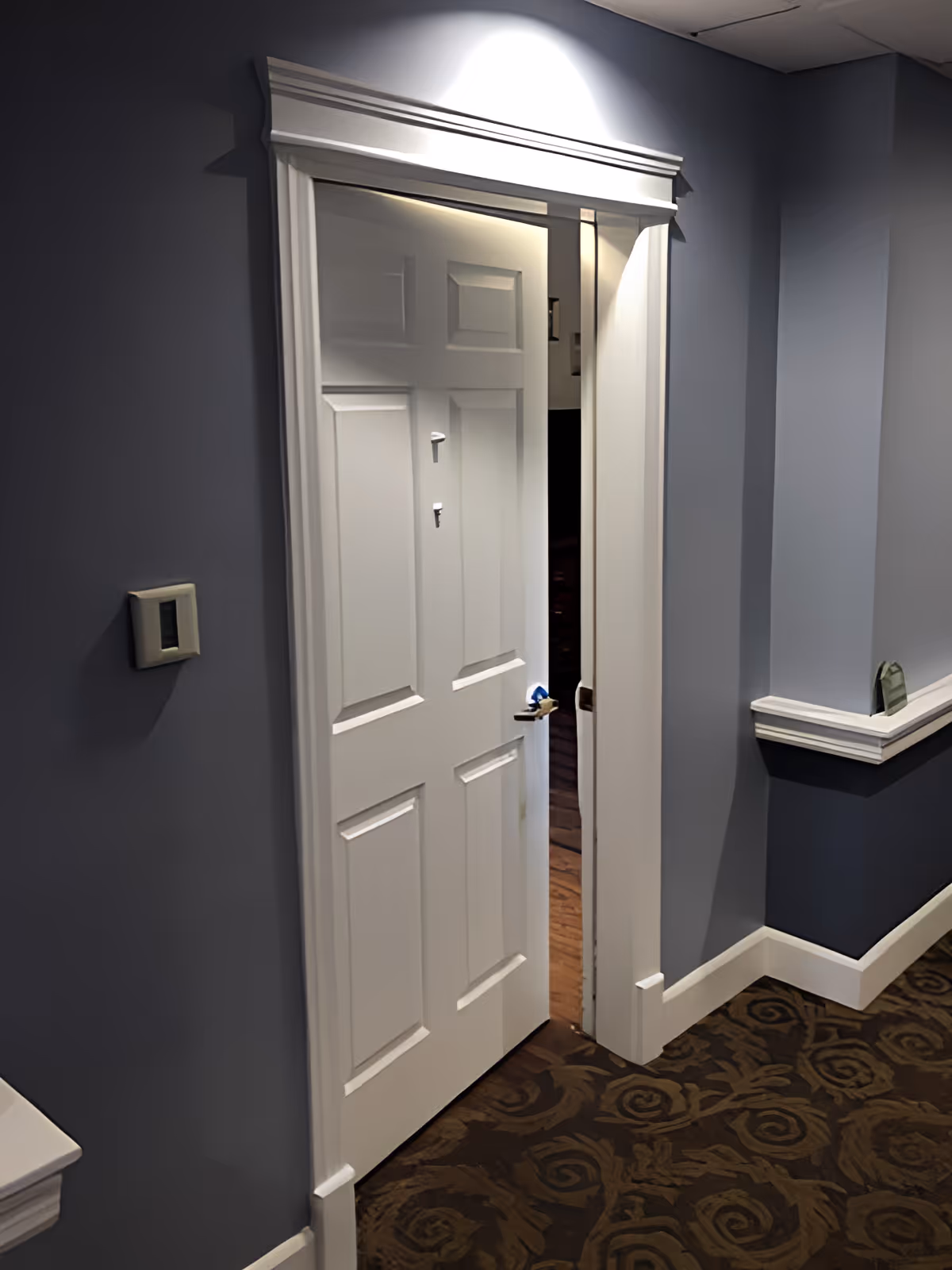 Partially open white door with decorative molding in a hallway with blue-gray walls and patterned carpet. A light switch is visible on the left wall and a small ledge with white trim is on the right wall.