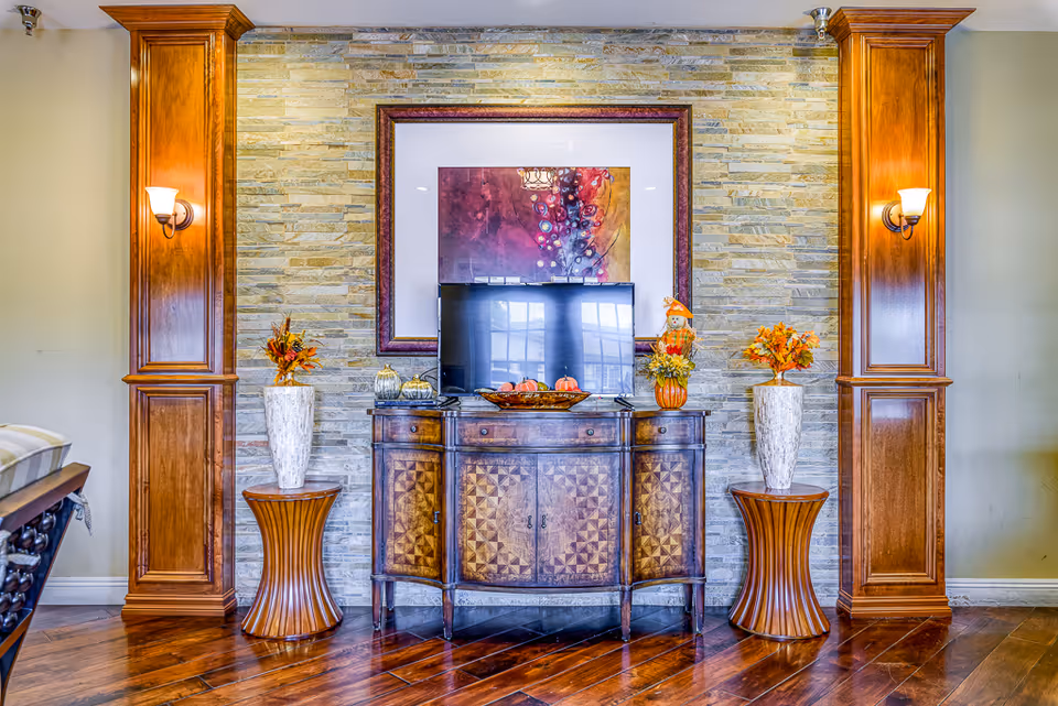 A cozy interior living room area with a wooden cabinet featuring intricate patterns, a flat-screen TV on top, and decorative items including a bowl of pumpkins and a small scarecrow figure. The background wall is made of light-colored stone tiles, flanked by two tall wooden columns with wall sconces. Two tall vases with autumn-themed floral arrangements sit on round wooden side tables on either side of the cabinet.