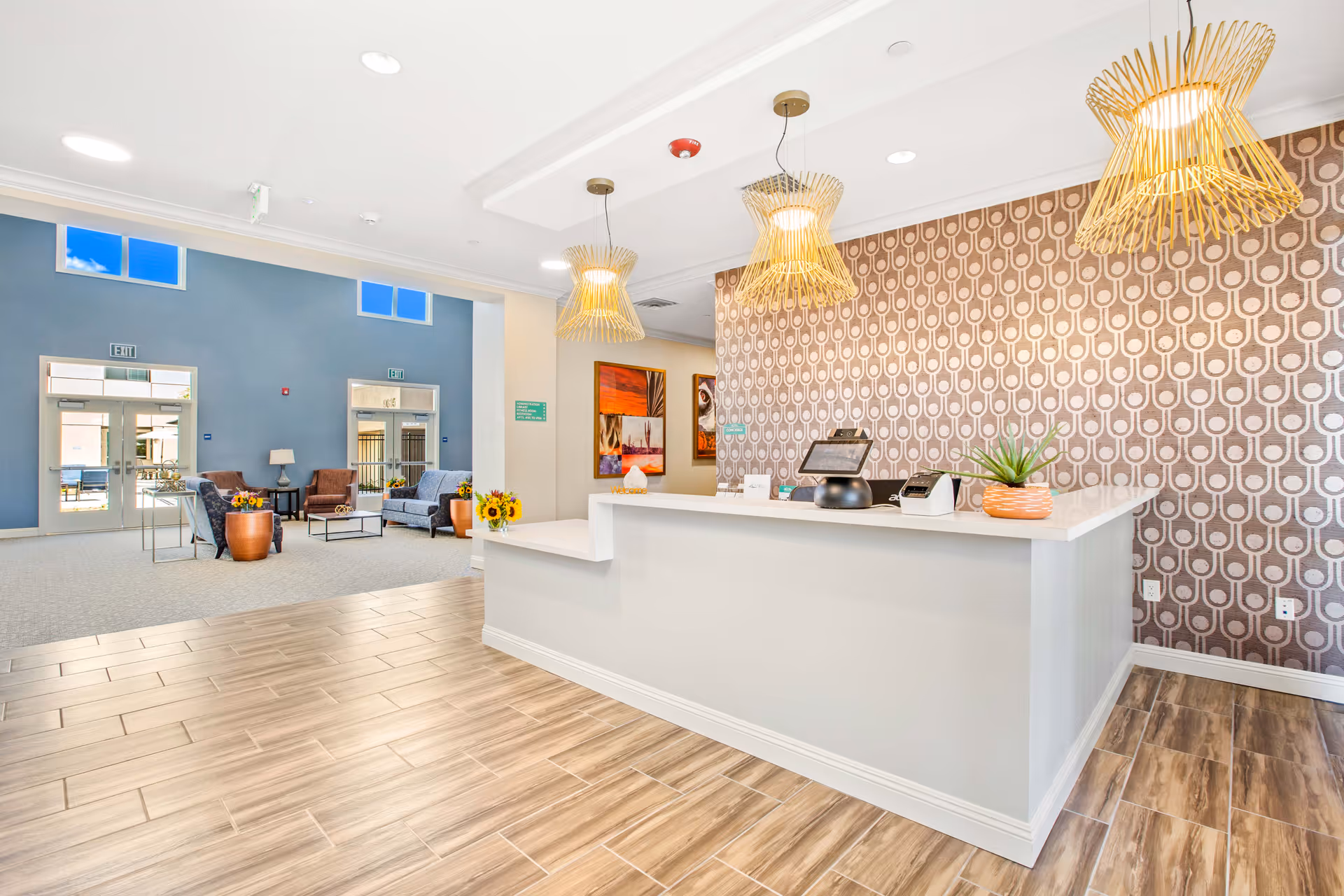 Bright and modern reception area of a senior living facility with a white reception desk, three decorative pendant lights, patterned wallpaper behind the desk, and a seating area with sofas and chairs near large windows and glass doors.