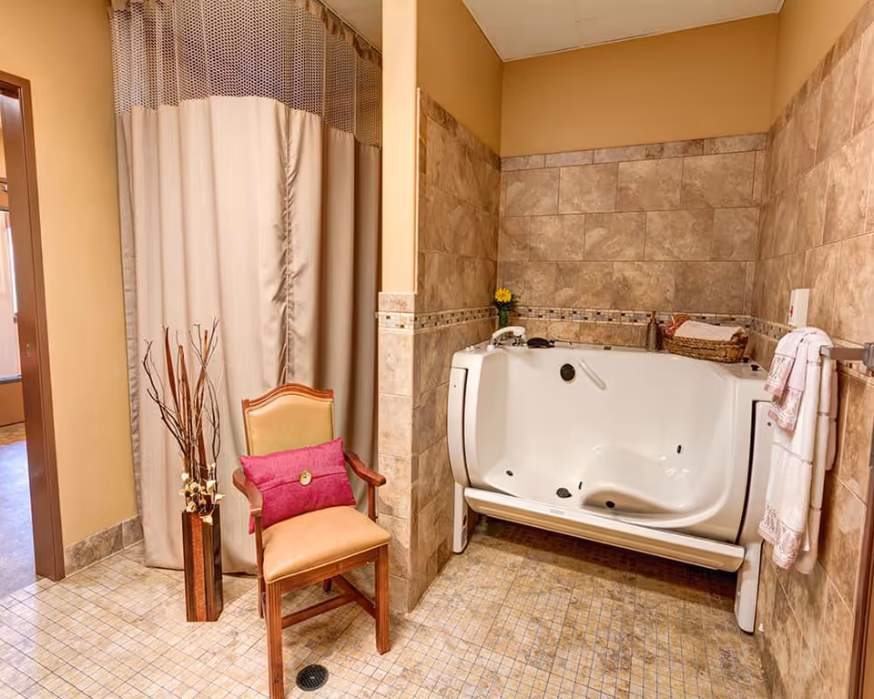 Accessible tiled bathroom featuring a white walk-in bathtub, an upholstered chair with a pink cushion, towels, and decorative accents.