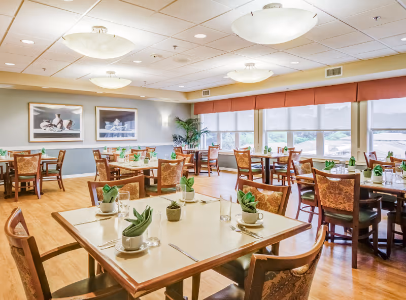 Bright dining room with multiple set tables and chairs, large windows, and ceiling lights.