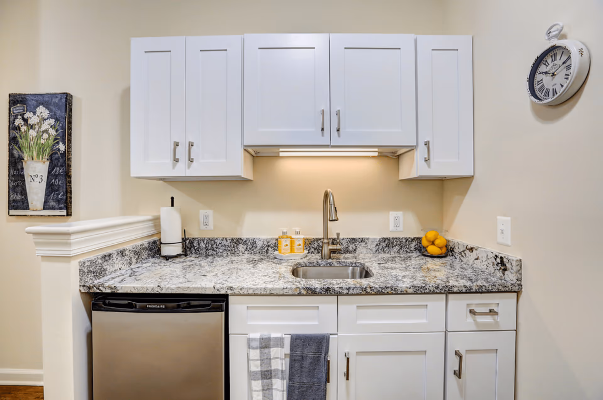 A small kitchen area with white cabinets, a granite countertop, a stainless steel sink with a modern faucet, a mini refrigerator, and a wall clock. There is a paper towel holder, a decorative painting of flowers, two hand towels hanging on the cabinet, and a bowl of lemons on the counter.
