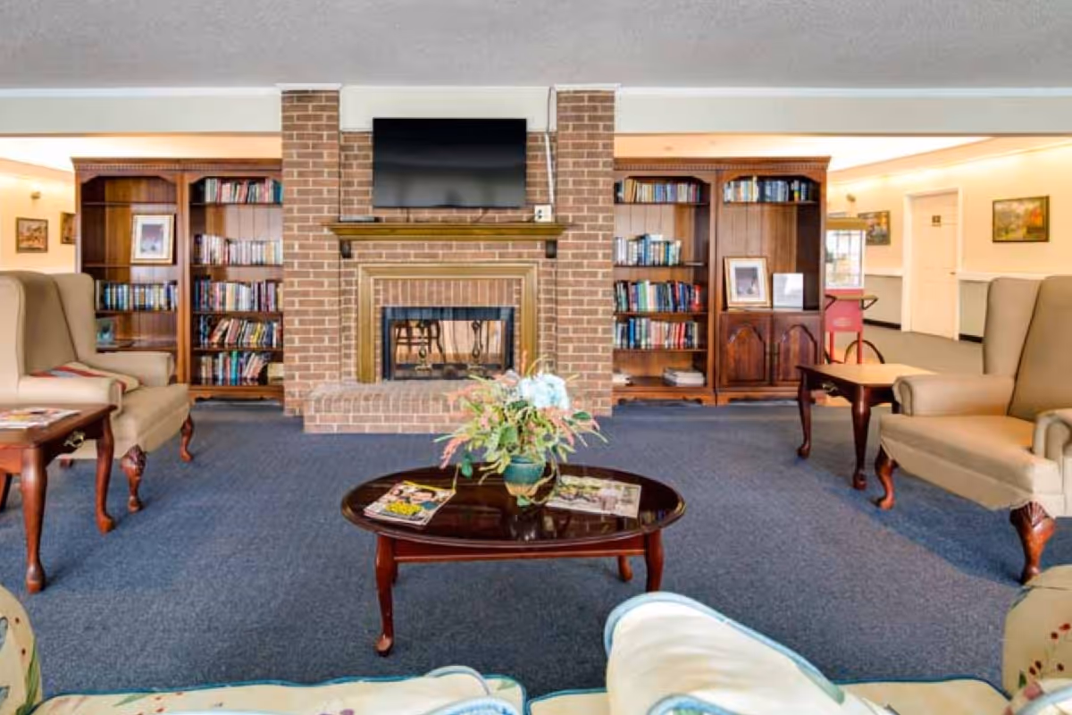 A cozy living room area with a brick fireplace in the center, a flat-screen TV mounted above it, two wooden bookshelves filled with books on either side, and two beige wingback chairs with wooden side tables. A wooden coffee table with a floral arrangement and magazines is in the foreground, and the room has blue carpeting and light-colored walls with framed artwork.