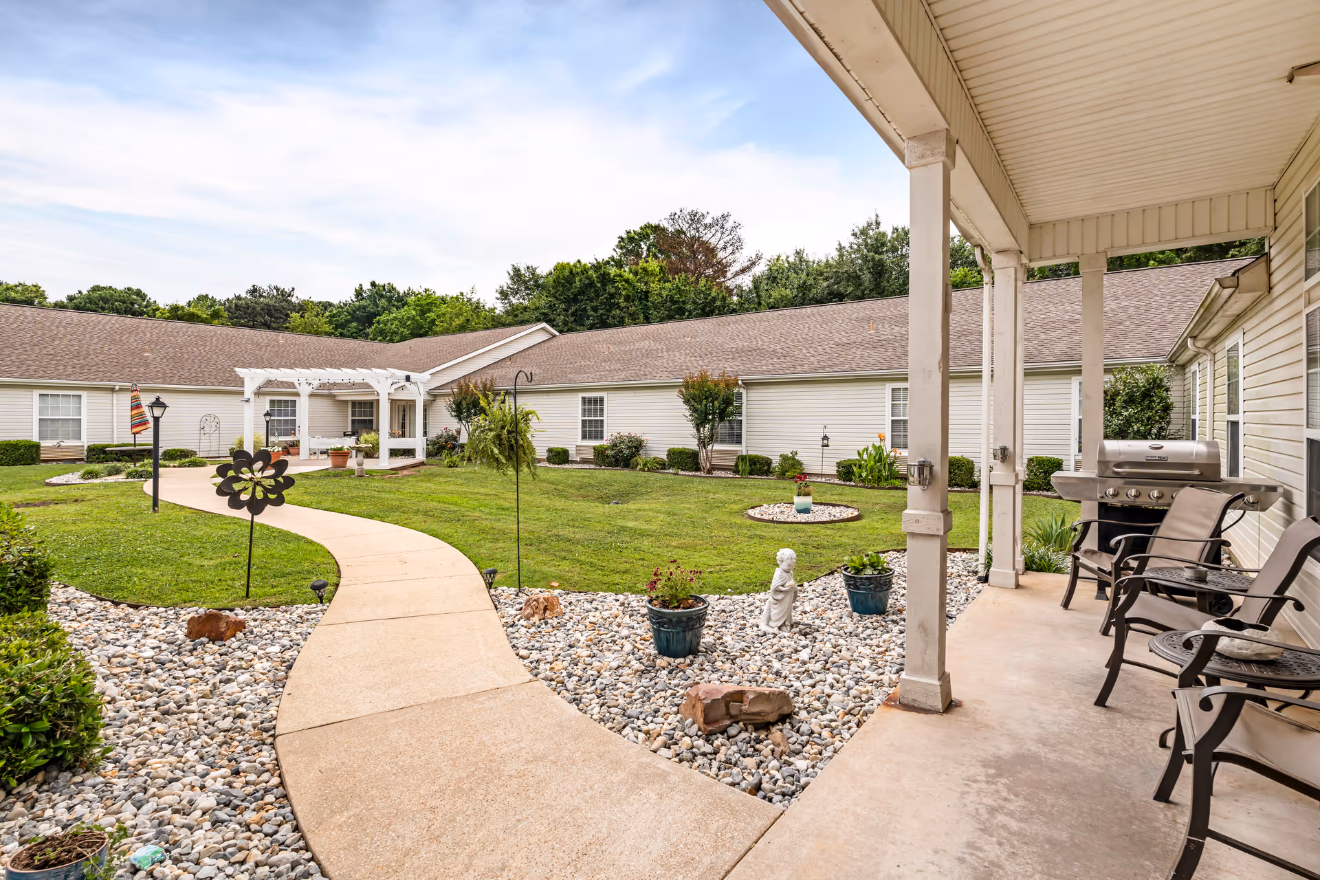 Outdoor courtyard area of a senior living facility with a curved concrete walkway, green lawn, decorative plants, and a white pergola. There is a covered patio with chairs and a barbecue grill on the right side. The building has beige siding and multiple windows, surrounded by trees and shrubs.