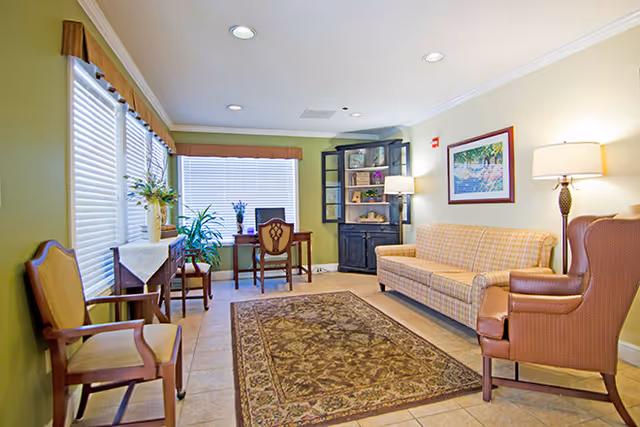 A cozy living room area with a patterned rug on tiled floor, a plaid sofa, a brown armchair, wooden chairs, a small table with a vase of flowers, a black cabinet with shelves and a lamp, and a framed picture on the wall. Large windows with blinds and valances let in natural light.