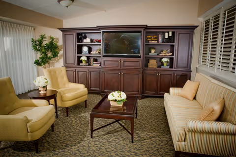 A cozy living room with a large dark wood entertainment center holding a flat-screen TV and decorative items. The room features a striped sofa with two pillows, two beige armchairs, a wooden coffee table with a flower arrangement, a side table with a flower vase, a potted plant, and carpeted flooring with a patterned design.