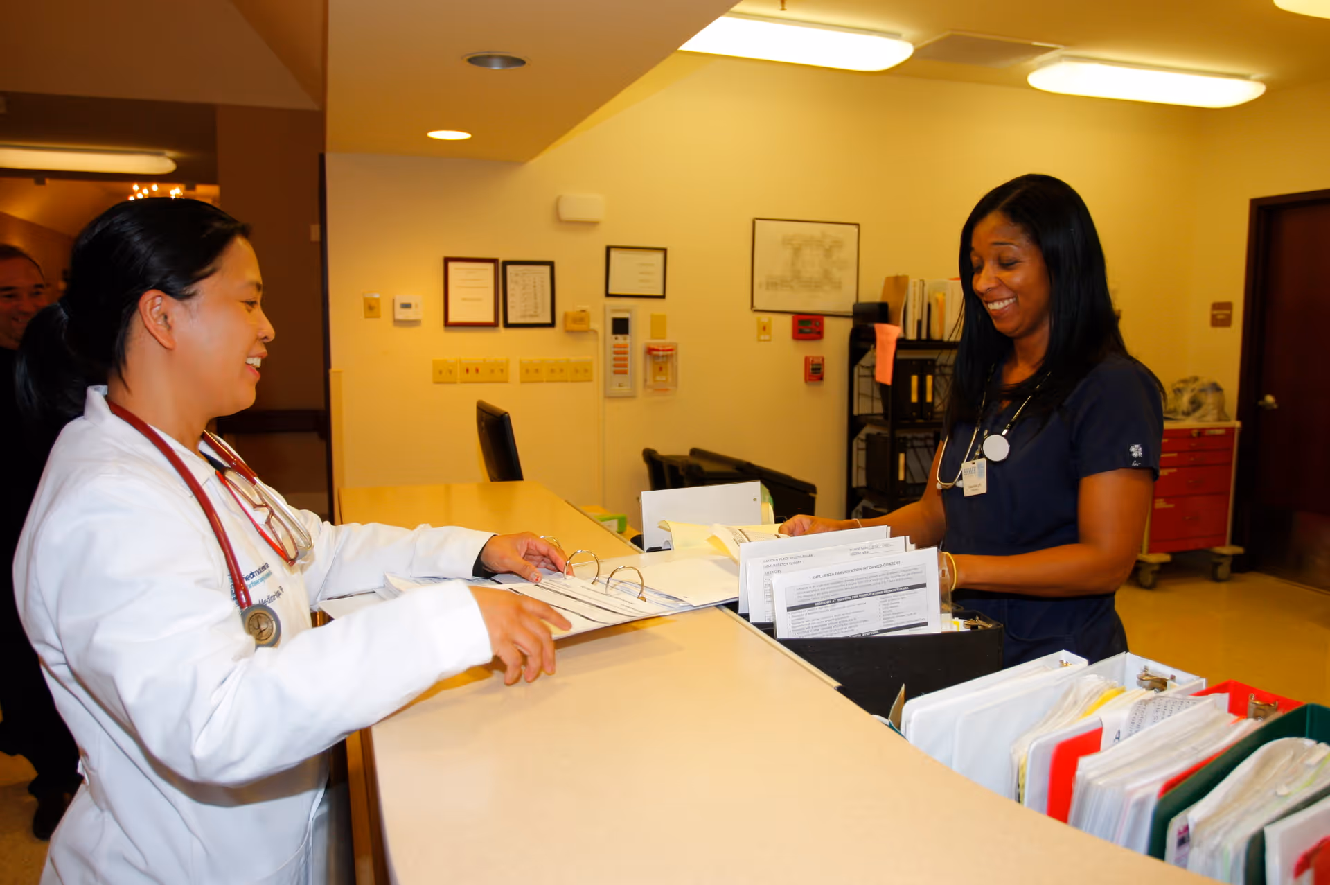 Two healthcare professionals, one in a white coat and the other in navy scrubs, are standing at a reception desk reviewing documents and smiling inside a medical facility.