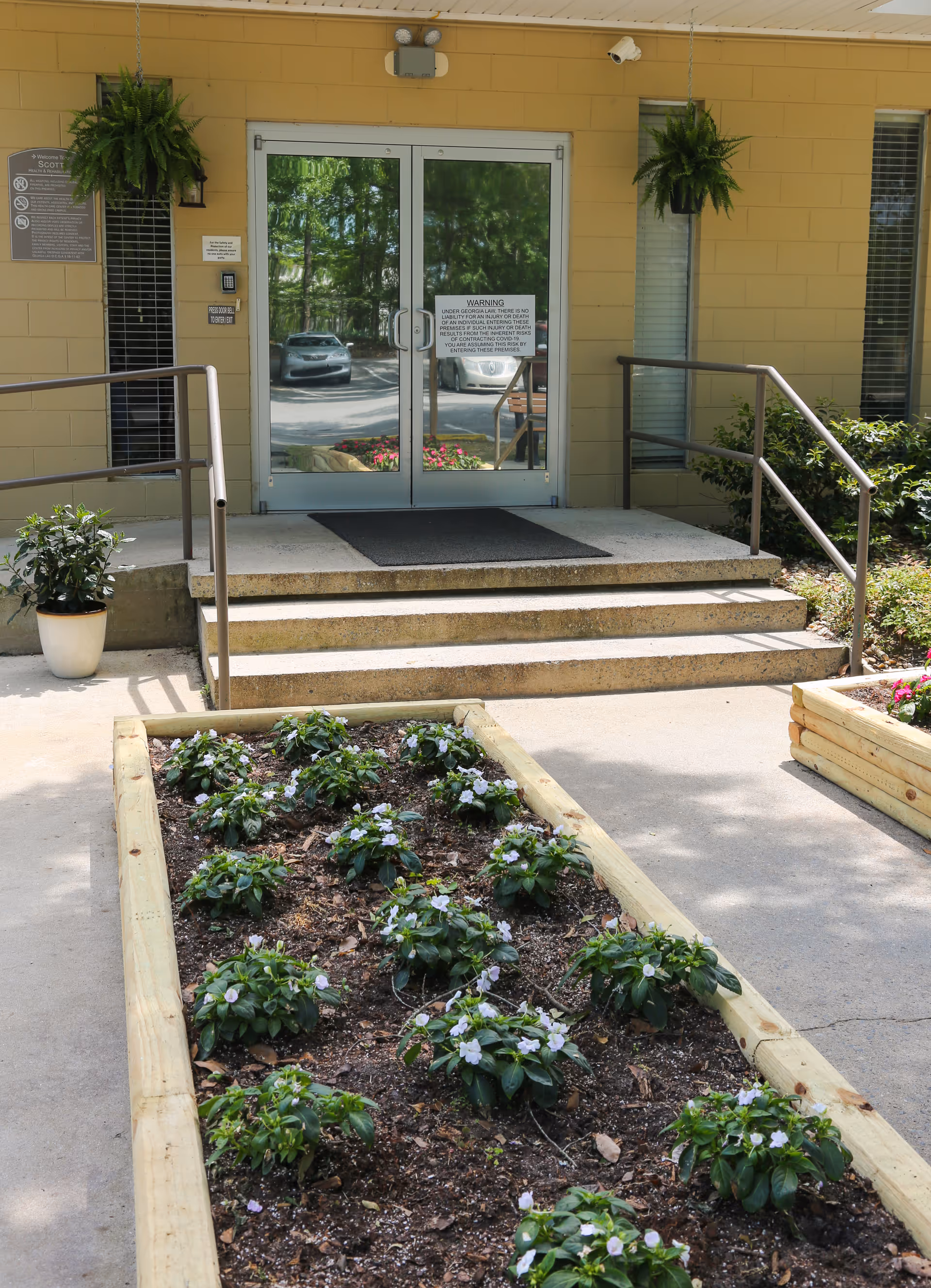 Entrance to Scott Health & Rehabilitation facility with glass double doors, concrete steps with metal railings, potted plants, and a raised garden bed with small flowering plants in front.