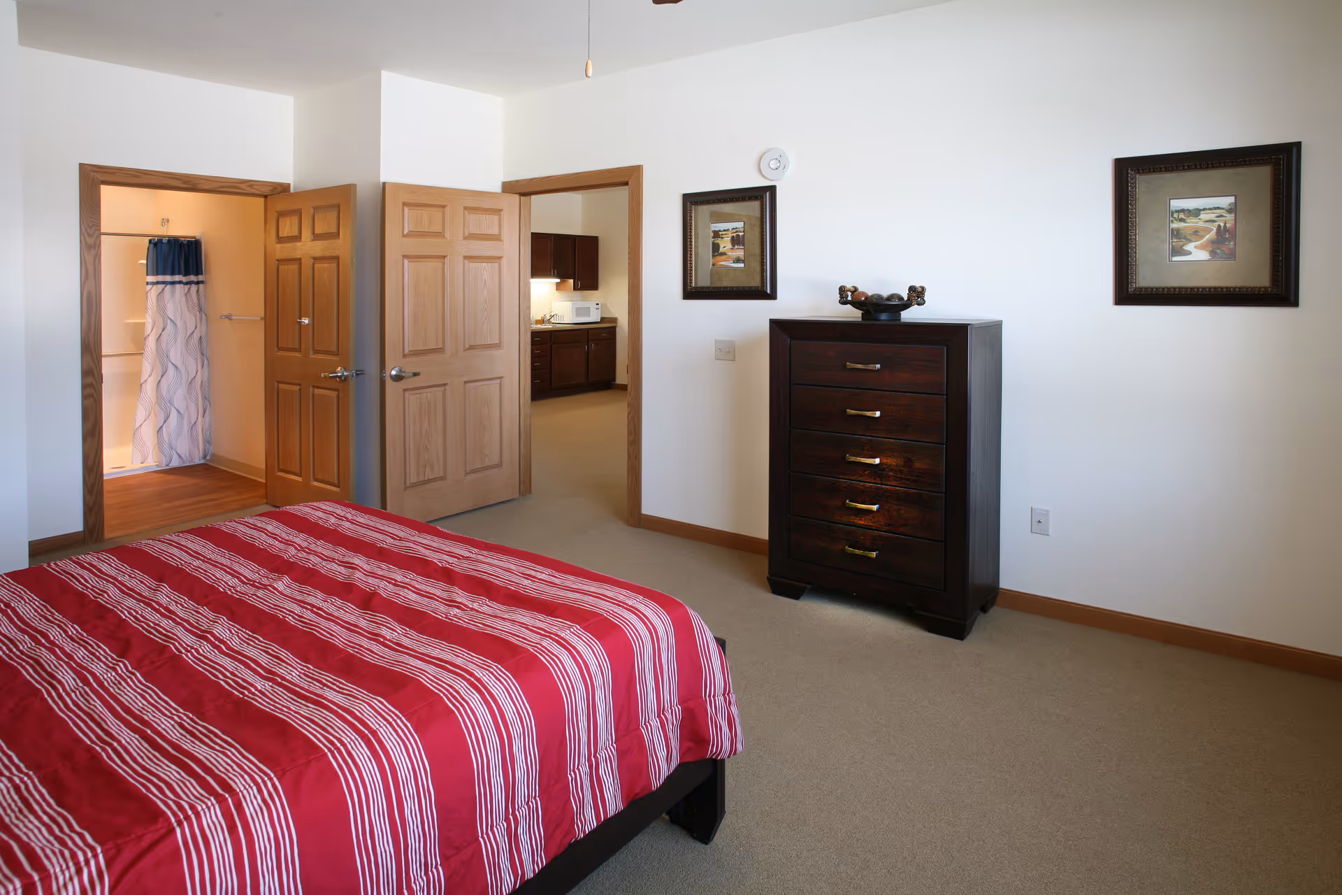 A bedroom with a bed covered in a red and white striped bedspread. There is a dark wooden chest of drawers against a white wall with two framed pictures above it. Two open wooden doors reveal a bathroom with a shower curtain and a kitchen area with cabinets and a microwave.