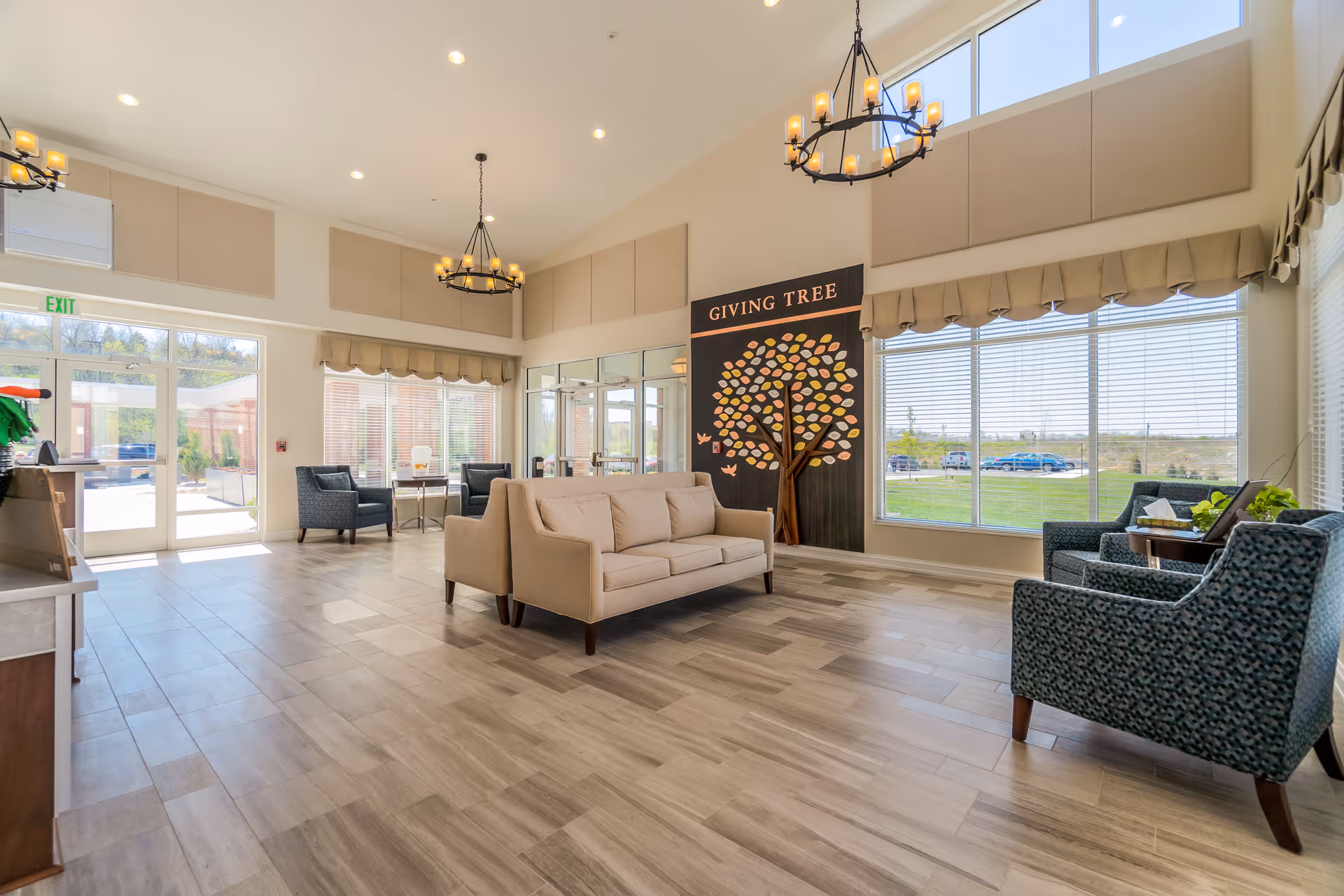 Bright, spacious senior living lobby with sofas and armchairs, chandeliers, large windows, and a 'Giving Tree' display.