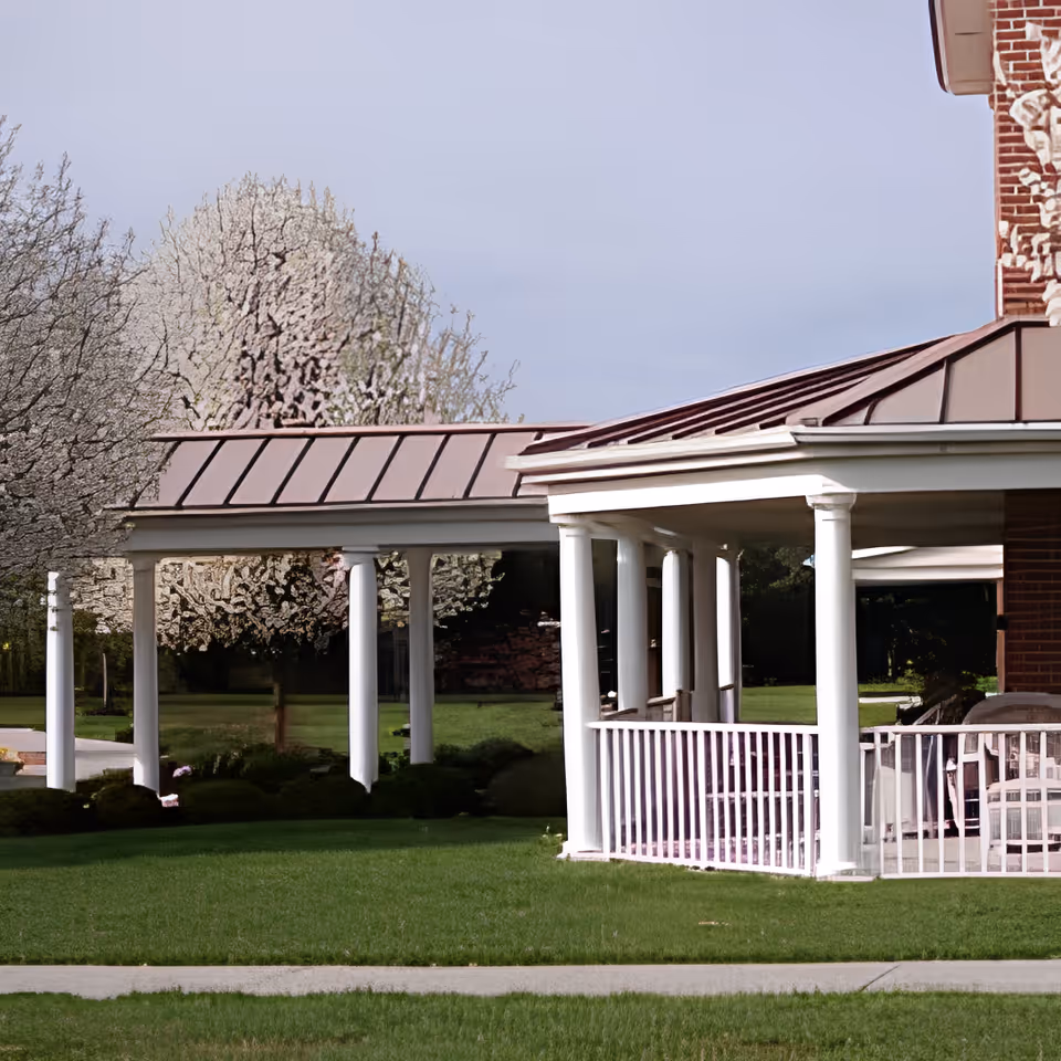 Covered white-columned porch and connecting walkway beside a brick building with a green lawn and blossoming trees.