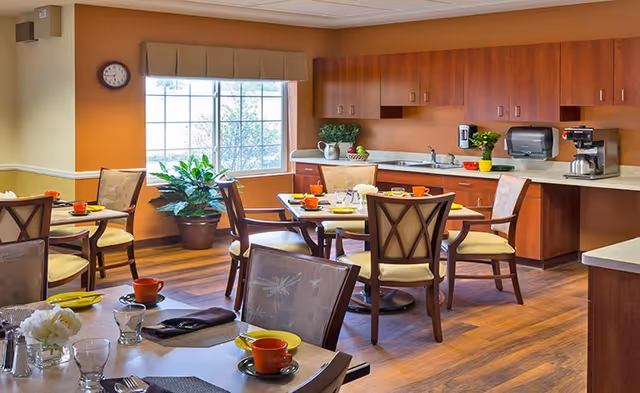 A bright dining room with wooden floors and orange walls. Several tables are set with plates, cups, and utensils. There are cushioned wooden chairs around the tables. A large window with a valance lets in natural light, and a potted plant is placed near the window. The back wall has wooden cabinets, a countertop with a coffee maker, a paper towel dispenser, and some decorative plants.