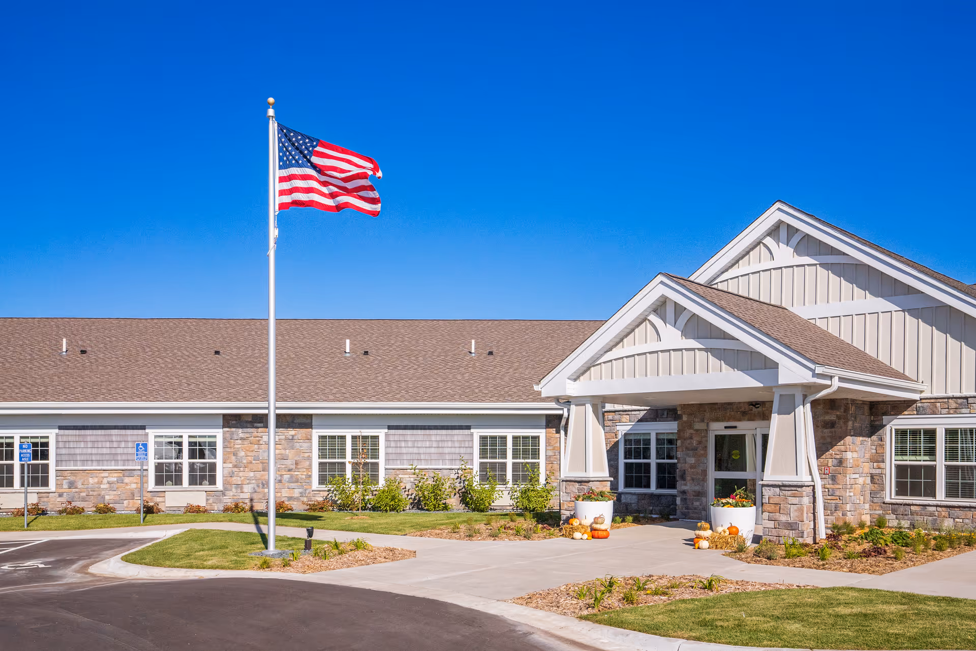 Front entrance of a single-story senior living building with an American flag on a flagpole and landscaped grounds.