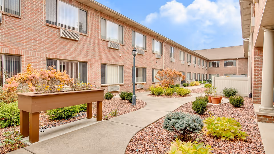 Outdoor courtyard area of a senior living facility with a concrete pathway, raised garden beds, various shrubs and plants, and a two-story brick building with multiple windows in the background under a partly cloudy sky.