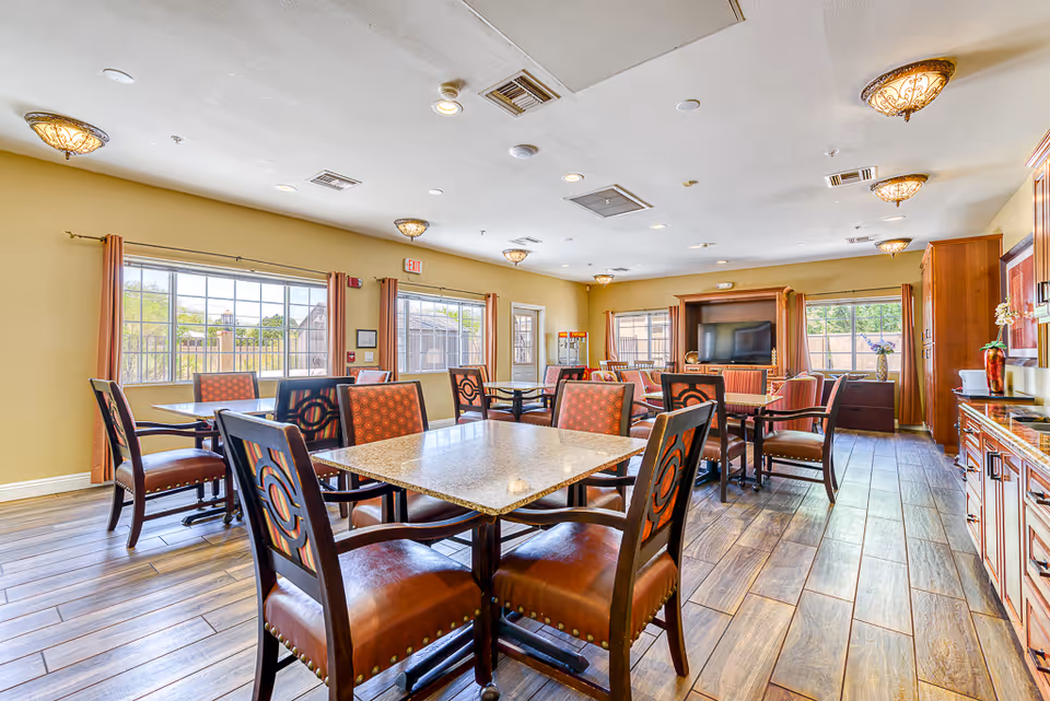 A bright and spacious common area with multiple tables and chairs arranged for seating. The room features large windows with curtains allowing natural light to enter, wood-look flooring, and warm yellow walls. There is a large flat-screen TV mounted on a wooden cabinet at the far end of the room, and decorative ceiling lights provide additional illumination.