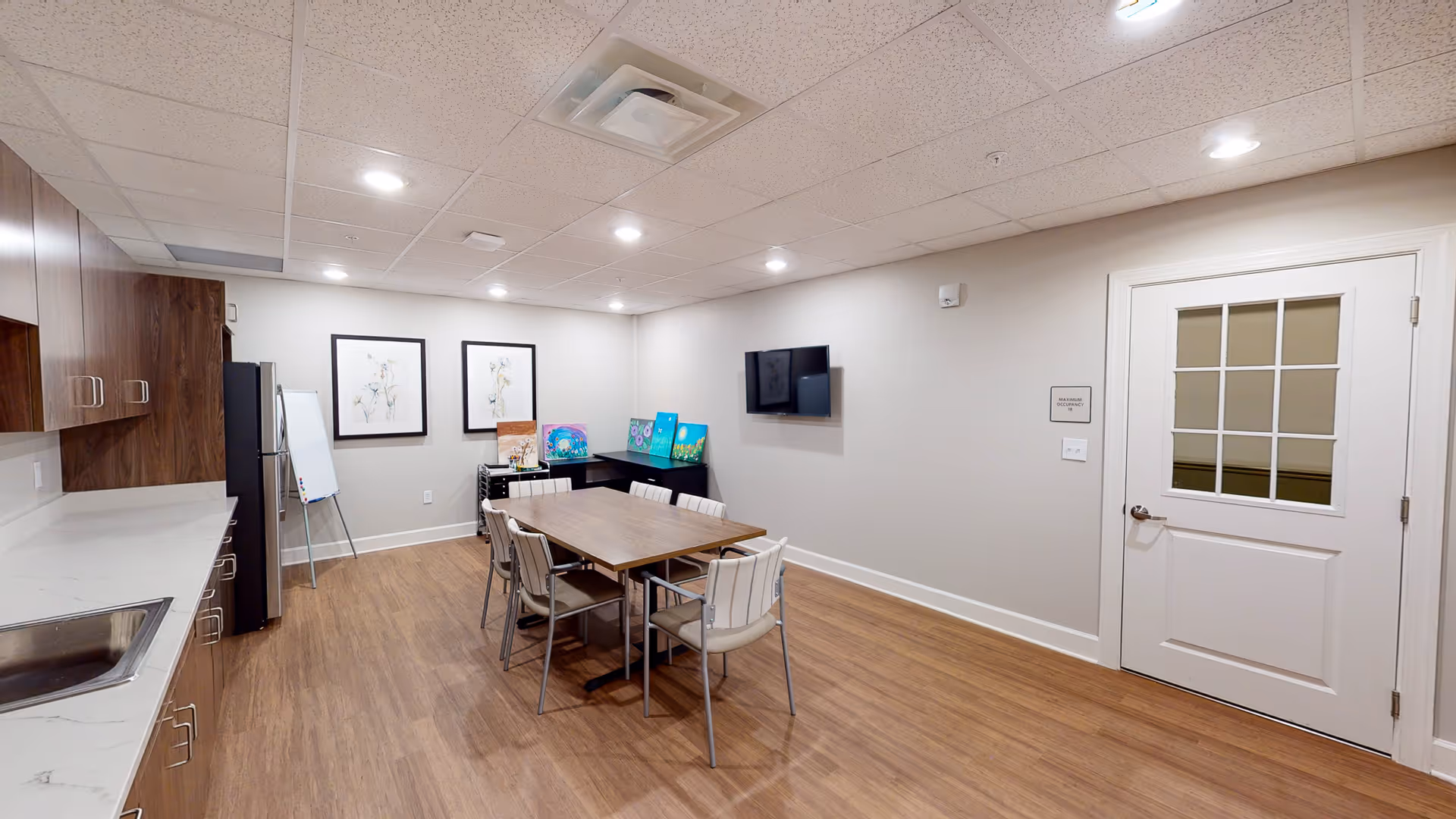 A well-lit room with a wooden table surrounded by six chairs in the center. To the left, there is a countertop with a sink and wooden cabinets above and below. A refrigerator and a whiteboard on an easel are against the far wall. On the right wall, a flat-screen TV is mounted above a sign that reads 'Maximum Occupancy 18'. Several colorful paintings rest on a black desk in the corner. The floor is wood, and the ceiling has recessed lighting and a ventilation unit.