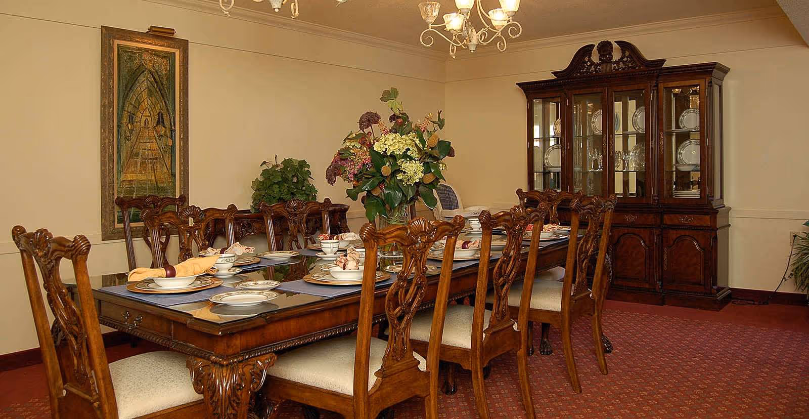 Formal dining room with a long ornate wooden table set with china, carved chairs, a floral centerpiece, chandelier, and a china cabinet.