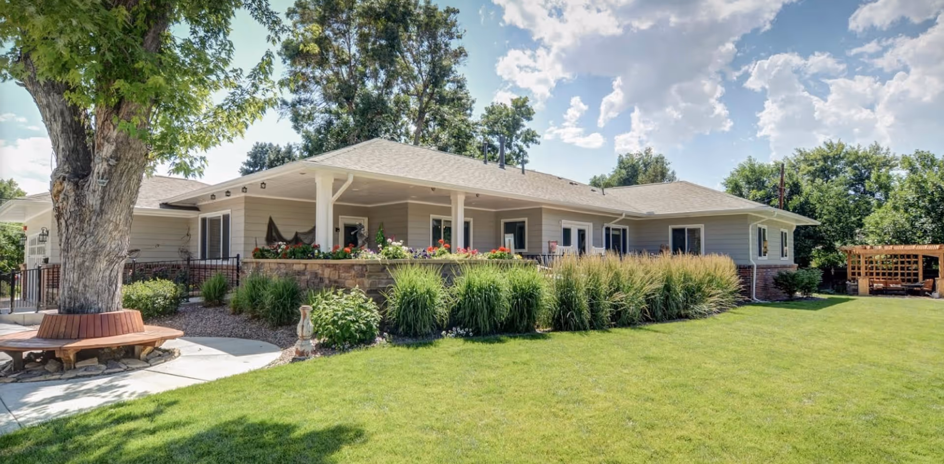 Exterior view of a single-story senior living facility building with a covered porch, surrounded by green grass, bushes, and trees under a partly cloudy sky. There is a large tree with a circular wooden bench around its base on the left side and a wooden pergola structure on the right side in the background.