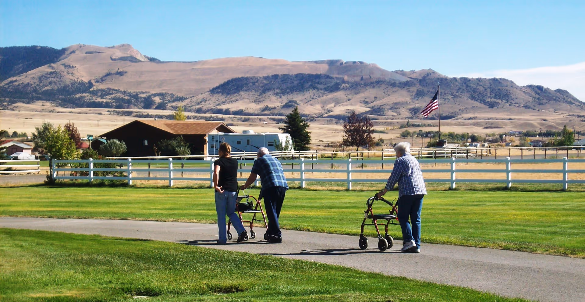 Two elderly individuals using walkers and a younger person walking together on a paved path in a grassy outdoor area with a white fence, houses, and mountains in the background under a clear blue sky.