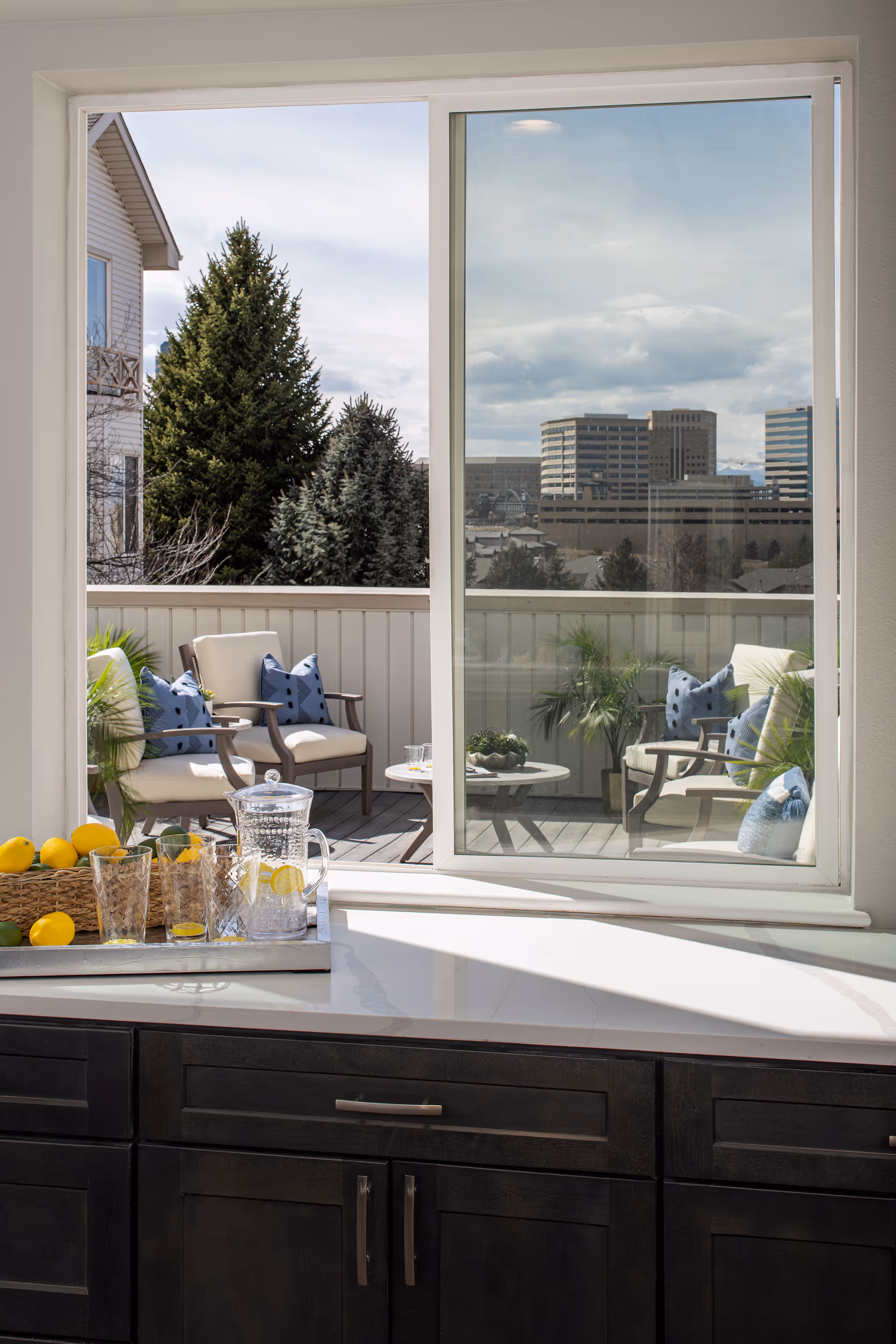 View from inside a kitchen looking out through a large window onto a balcony with outdoor seating. The balcony has cushioned chairs with blue pillows, a small round table, and some potted plants. In the foreground, a kitchen counter holds a tray with a glass pitcher of lemonade, glasses, and a basket of lemons. In the background, there are trees and city buildings under a partly cloudy sky.