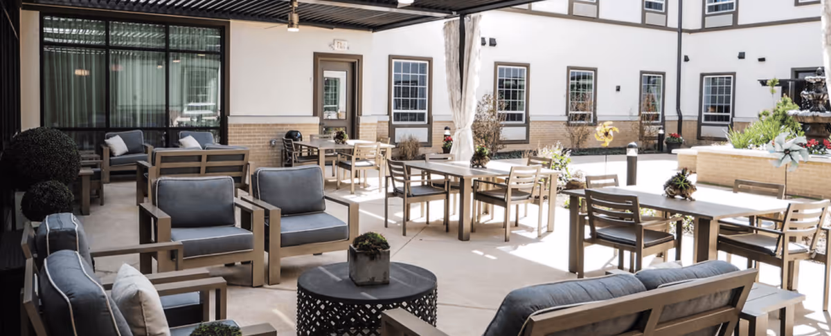 Outdoor patio area with multiple seating arrangements including cushioned chairs and sofas, tables with chairs, potted plants, and a water fountain, surrounded by a building with windows.