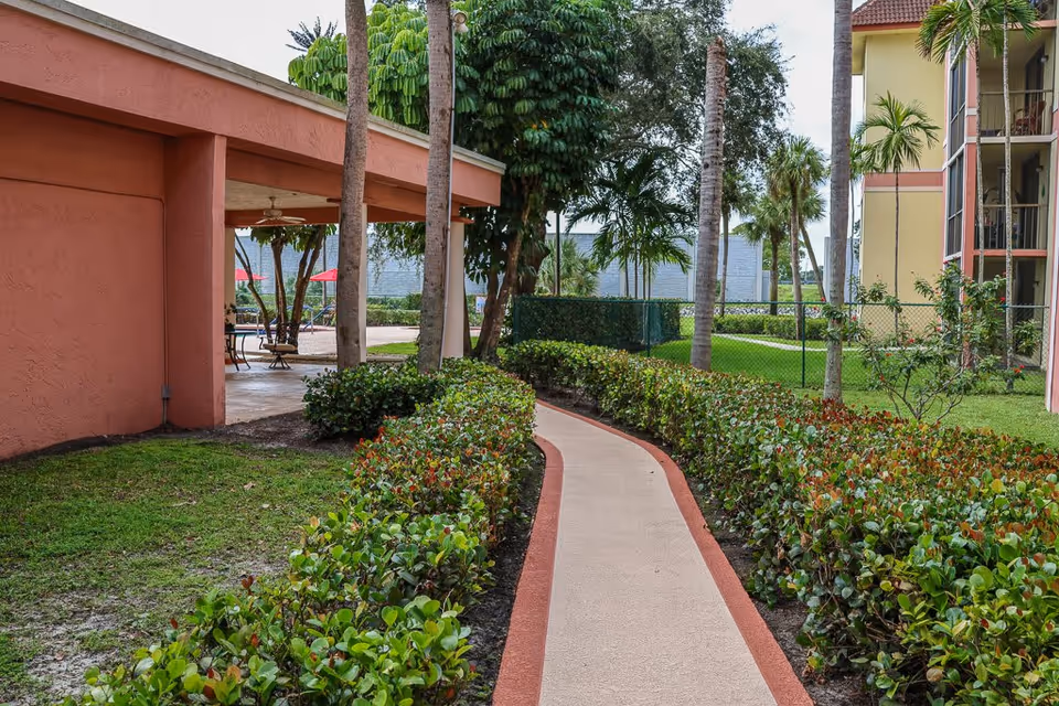 A paved walkway bordered by green shrubs and palm trees leads through a landscaped outdoor area between buildings with peach and yellow walls. There is a covered patio area with tables and chairs visible on the left side, and a chain-link fence separates the walkway from a grassy area with more palm trees.