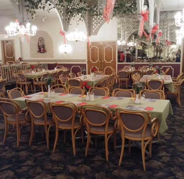 A dining room with multiple tables covered in light green tablecloths, each set with red napkins, menus, and small flower arrangements. The room features wooden chairs with cushioned seats, chandeliers hanging from the ceiling, decorative arches with greenery, and a large mirror on one wall reflecting the room.