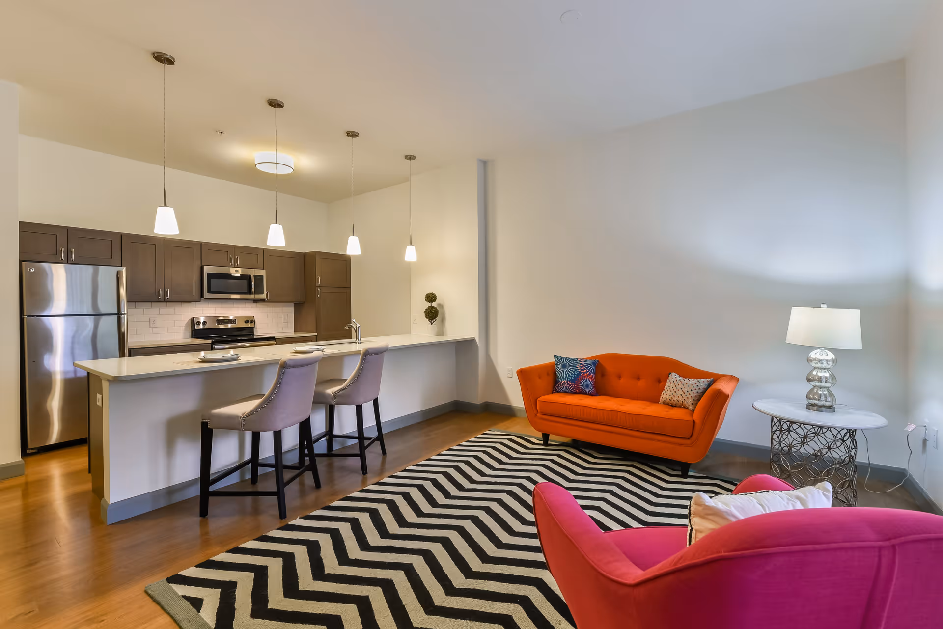 A modern open-concept living area featuring a kitchen with stainless steel appliances, dark wood cabinets, and a white countertop island with two beige bar stools. The living space includes a bright orange sofa, a pink armchair, a black and white zigzag patterned rug, and a round side table with a silver lamp. The walls are painted white and the floor is wooden.