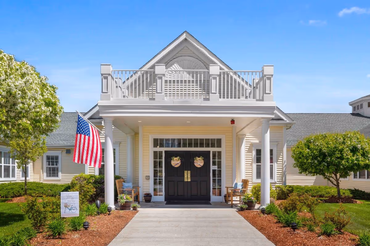 Front entrance of a senior living facility with a covered porch supported by white columns, two wooden chairs with cushions on either side of the black double doors, an American flag on the left, and well-maintained landscaping with green bushes and trees under a clear blue sky.