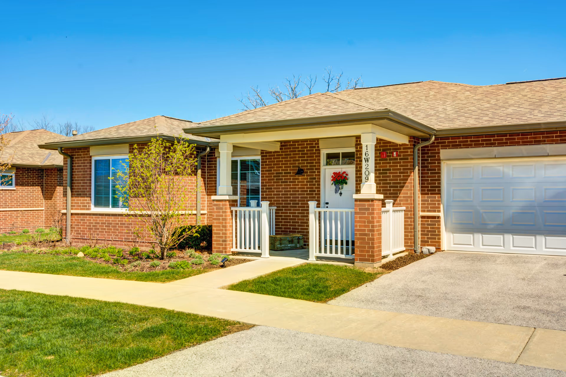Exterior view of a single-story brick senior living unit with a small covered porch, white railing, a white front door decorated with a red flower wreath, and an attached garage. The unit is surrounded by a well-maintained lawn and small shrubs under a clear blue sky.