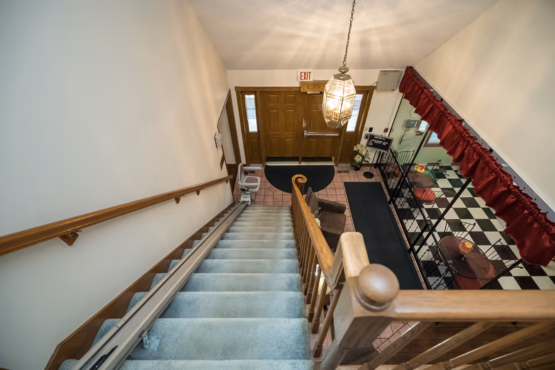 View looking down a carpeted staircase with wooden handrails into an entrance area with double wooden doors, a hanging light fixture, a chair lift on the stairs, a brown armchair, and a seating area with round tables and chairs on a black and white checkered floor with red curtains.