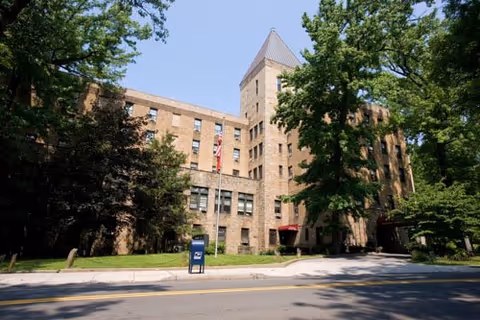 Exterior view of a multi-story brick building with a tower-like structure in the center, surrounded by trees and greenery under a clear blue sky. A mailbox and a flagpole with an American flag are visible near the sidewalk in front of the building.